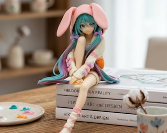 May include: A collectible anime-style figurine of a character with long teal hair, wearing a light pink bunny-eared hood, a cream-colored sweater with pastel accents, and matching striped socks, sitting atop a stack of three hardcover books.