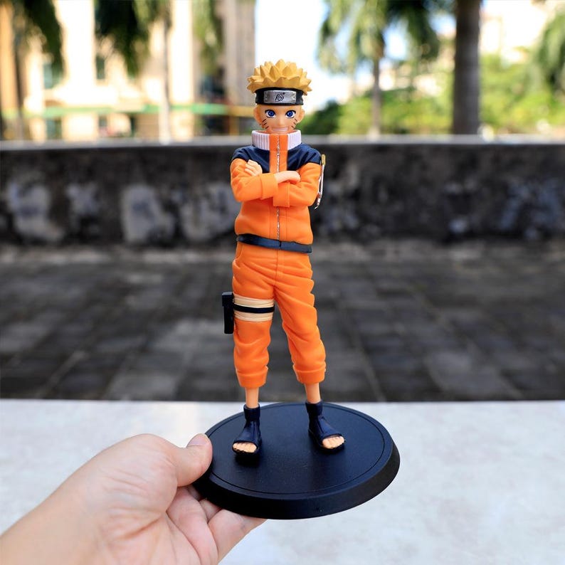 May include: A standing anime character figurine with spiky blonde hair, wearing a bright orange jumpsuit with a dark blue collar and accents. The figure has blue eyes, a headband with a metal plate, and is mounted on a round black base.
