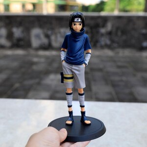 May include: A Sasuke Uchiha anime character figurine featuring black hair, a dark blue high-neck shirt, light gray shorts, and leg wraps. The figure stands on a circular black base and wears a headband with a leaf symbol on the forehead.