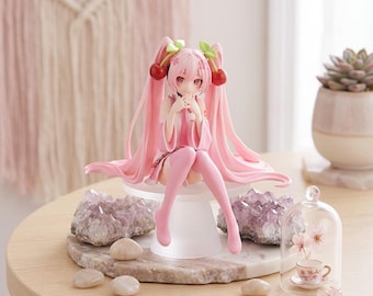May include: A pink-haired anime-style figurine with long twin tails adorned with cherry hair accessories. The character wears a light pink outfit and sits on a white pedestal. The figure features large eyes and is posed with hands near the face.