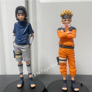 May include: Two anime character figurines standing on black circular bases. The left figure has black hair, a dark blue shirt, and gray shorts. The right figure has spiky blonde hair, an orange jumpsuit, and arms crossed. Text reads TAYI Toys Sro.