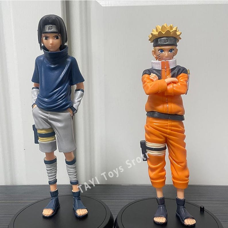 May include: Two anime character figurines standing on black circular bases. The left figure has black hair, a dark blue shirt, and gray shorts. The right figure has spiky blonde hair, an orange jumpsuit, and arms crossed. Text reads TAYI Toys Sro.