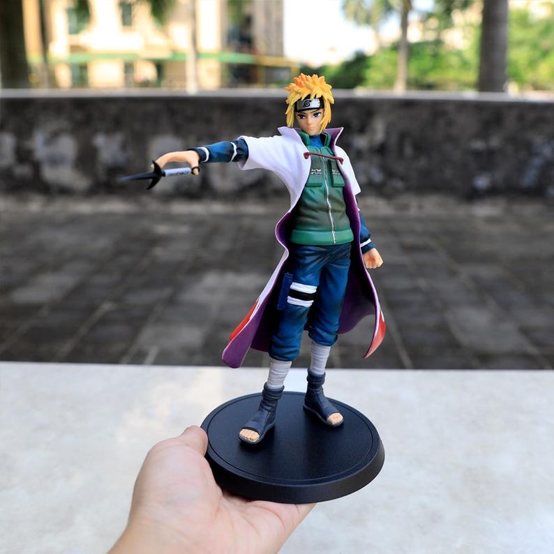 May include: A Minato Namikaze anime figurine featuring spiky yellow hair, a white coat with red flame patterns, a green vest, and blue pants. The character holds a black kunai weapon in one hand and stands on a circular black base.