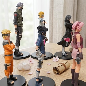 May include: A collection of six anime character figurines standing on circular black bases. The figures include characters with spiky blonde, black, silver, and pink hair, wearing various outfits like orange jumpsuits, dark cloaks, and tactical gear.