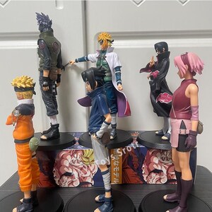 May include: A collection of five anime character figurines standing on black circular bases. The figures include characters with spiky yellow, black, and pink hair, wearing outfits in orange, dark green, white, blue, and black, with red cloud patterns.