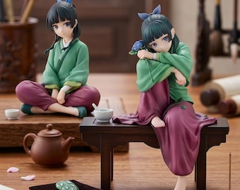 May include: Two anime-style figurines depicting a character with dark hair in a green tunic and maroon pants. One figure sits cross-legged on a wooden platform, while the other sits on a small wooden bench holding a blue flower, surrounded by tea items.