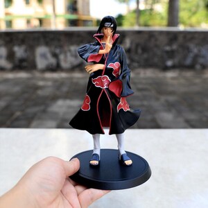 May include: A plastic anime character figurine featuring a person with black hair wearing a long black cloak with red cloud patterns and a red interior lining. The figure stands on a circular black base, wearing white pants and dark sandals.