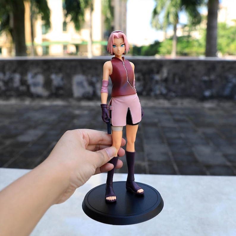 May include: A standing anime character figurine with short pink hair, wearing a sleeveless dark red zip-up top, a light pink skirt, purple arm warmers, and dark purple calf-high boots, mounted on a circular black base.