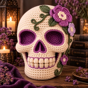 Puede incluir: Una calavera de ganchillo color crema con cuencas oculares y cavidad nasal de color morado oscuro. La calavera está decorada con enredaderas de ganchillo verde oscuro y varias flores moradas y lila claro con centros crema.