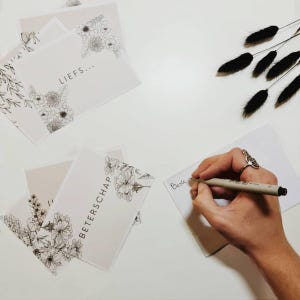 Op de afbeelding: Een verzameling witte rechthoekige wenskaarten met zwarte lijntekeningen van bloemen. Eén kaart bevat de tekst "LIEF..." en een andere "BETERSCHAP". Een hand schrijft "Beste" op een lege kaart met een zwarte fineliner.