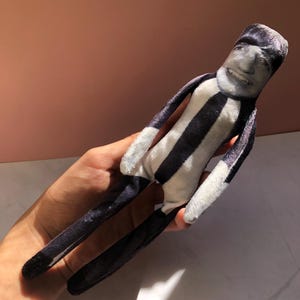May include: A plush doll featuring a printed human face with a smiling expression. The body is made of soft fabric with vertical black and white stripes. The doll has long, slender arms and legs, and a cylindrical head shape with a realistic photographic print.