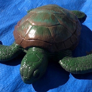 May include: A sea turtle figurine featuring a glossy, textured green head and flippers with a rounded shell painted in shades of brown and green. The surface of the turtle has a smooth, reflective finish, highlighting the detailed scale patterns.