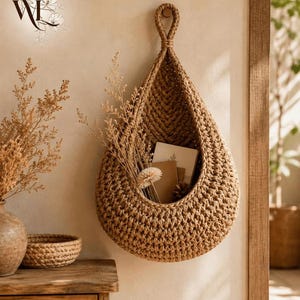 May include: A teardrop-shaped wall hanging basket made of thick, tan-colored woven cord. The basket features a textured, braided pattern and a loop at the top for mounting. Inside the basket are several small books and dried floral stems.