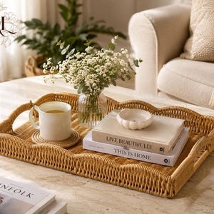 Puede incluir: Una bandeja rectangular de ratán trenzado con bordes festoneados y asas laterales. Dentro, una taza de cerámica blanca descansa sobre un pequeño posavasos, junto a un jarrón de cristal transparente con flores blancas, un cuenco blanco pequeño y dos libros apilados titulados Live Beautifully y This Is Home.