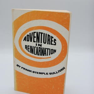 May include: A vintage book titled "Adventures in Reincarnation" by Frank Stemple Sullivan. The book cover features an orange and white design with the title in black lettering. The book is standing upright against a white background.