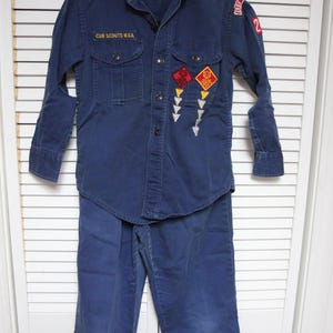 Op de afbeelding: Een vintage Cub Scouts B.S.A. uniformset in marineblauw, inclusief een shirt met lange mouwen met borstzakken en patches, en een bijpassende broek. Het shirt heeft de tekst "CUB SCOUTS B.S.A."