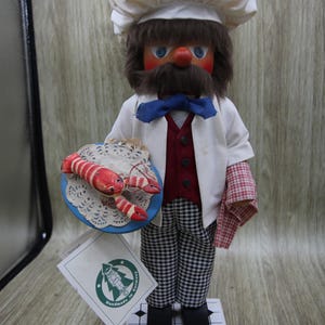 Peut inclure: Figurine de chef en bois, coiffé d'une toque et d'une veste blanches, avec un pantalon à carreaux. La figurine tient une assiette avec un homard rouge et blanc. Le chef a une barbe et une moustache brunes, et un nœud papillon bleu. Socle blanc.
