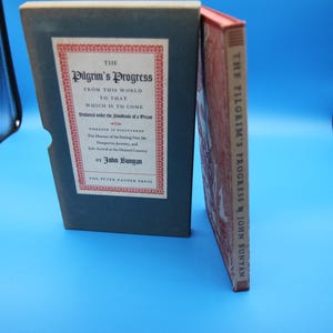 Może przedstawiać: Zabytkowy egzemplarz "Wędrówki Pielgrzyma" autorstwa Johna Bunyana. Książka znajduje się w ciemnoniebieskim etui z czerwoną i białą etykietą. Książka ma czerwony grzbiet i zniszczoną okładkę, z widocznym tytułem.