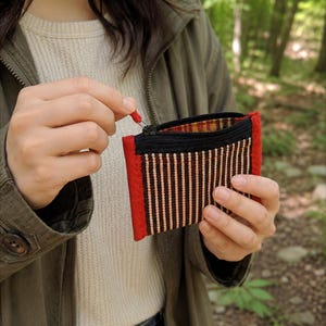 May include: A small rectangular zipped pouch featuring a woven pattern of vertical black and tan stripes. The pouch is framed by bright red fabric borders on the sides and top, with a black zip closure and a small red pull tab attached.
