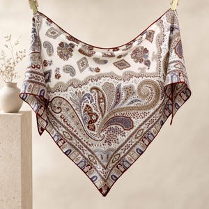 Könnte beinhalten: Ein quadratisches Tuch mit cremefarbener Basis und einem komplexen Paisley-Muster in Braun-, Blau- und Dunkelrottönen. Der Stoff hat einen dunkelroten Rand an den Kanten und ist mit zwei Holzklammern vor einem neutralen Hintergrund aufgehängt.