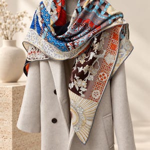Peut inclure: Un grand foulard carré présentant un motif complexe et multicolore composé de fleurs, de formes géométriques et de rayons de soleil dans des tons de bleu, rouge, marron et crème, drapé sur un manteau beige clair sur un cintre en bois.