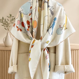 May include: A square scarf with a cream background featuring a colorful print of stylized animals, including a horse and birds, alongside branches with blue and orange fruit and leaves. The scarf is draped over a plain, light-colored coat on a hanger.