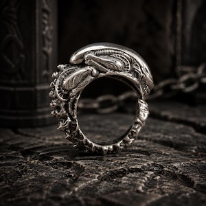 May include: A silver-toned ring featuring a highly detailed, sculpted creature design with an elongated, smooth head and textured, segmented body elements wrapping around the band. The metal surface has a dark, oxidized finish in the crevices.