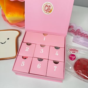 May include: A pink rectangular box with a hinged lid featuring a circular logo with the letters CS. The interior contains seven numbered pink drawers arranged in a grid, with one large drawer labeled 1 and six smaller drawers labeled 2 through 7.