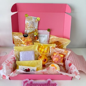 May include: A bright pink cardboard box filled with various individually packaged squishy toys and craft items. The contents include small plastic bags with colorful food-themed designs, a purple block, and a sign reading Cafescoopshop.