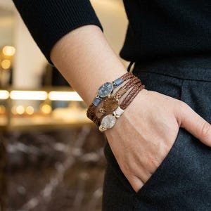 May include: A multi-strand wrap bracelet featuring three distinct oval-shaped stone charms in shades of blue, brown, and white. The bracelet is constructed with braided brown leather cords and metallic accents, designed to wrap around the wrist.