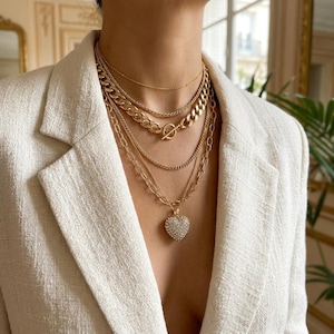 May include: A layered gold-tone necklace set featuring a delicate chain, a medium-weight curb chain with a toggle clasp, and a longer chain with a heart-shaped pendant encrusted with small clear stones, worn over a textured cream-colored blazer.