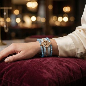 Puede incluir: Una pulsera de varias hebras con cuentas de color azul claro y un dije circular dorado en el centro que representa un árbol. La pulsera está superpuesta con una banda texturizada de color azul claro y un cierre dorado.
