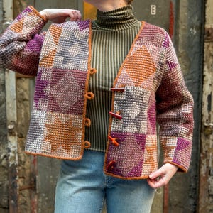 Puede incluir: Una chaqueta de ganchillo de manga larga con un patrón de patchwork de formas geométricas en tonos naranja, morado y gris. La chaqueta tiene un escote en V, mangas largas y un cierre frontal asegurado con cuatro botones de palanca oscuros.