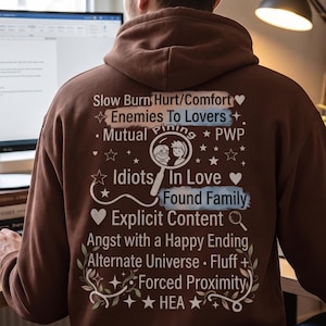 May include: A brown hooded sweatshirt featuring a back graphic with white and blue text listing various literary tropes including Slow Burn, Enemies To Lovers, Mutual Pining, PWP, Idiots In Love, Found Family, Explicit Content, Angst, and HEA.