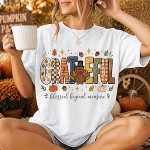 May include: A white short-sleeve t-shirt featuring the word GRATEFUL in large, multi-patterned letters, with a turkey wearing a hat replacing the letter A. Below, the text reads blessed beyond measure, surrounded by pumpkins and acorns.