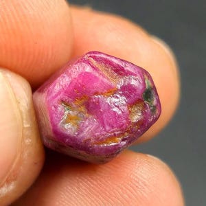 May include: A raw, unpolished ruby gemstone with a vibrant magenta and pink hue. The stone features a hexagonal shape with visible natural surface textures, including small brown inclusions and crystalline facets across its surface.