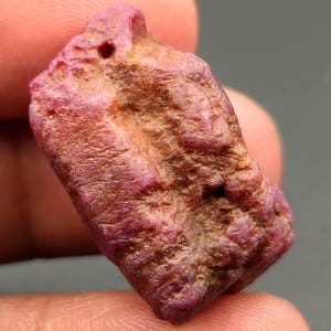 May include: A raw, unpolished ruby gemstone with a rough, textured surface. The stone displays a deep magenta and reddish-pink coloration with patches of earthy brown mineral deposits across its irregular, blocky, and elongated crystalline form.