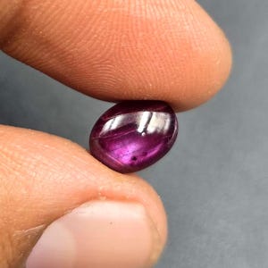 May include: A deep magenta oval cabochon gemstone with a smooth, polished surface and a distinct light reflection across the top. The stone displays a rich, saturated purple-red hue with subtle internal inclusions visible within the gem.
