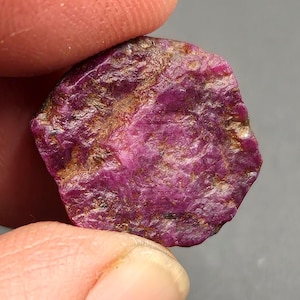 Peut inclure: Une pierre précieuse rubis brute de forme hexagonale avec une palette de couleurs magenta et cramoisi profond. La surface de la pierre présente une texture naturelle non polie avec des inclusions cristallines et des dépôts minéraux bruns.