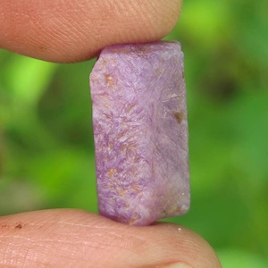 May include: A raw, rectangular-shaped gemstone with a light purple and lavender hue. The surface of the stone displays a fibrous, crystalline texture with subtle variations in colour and small, embedded brownish mineral inclusions throughout.