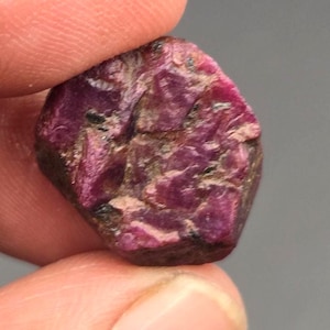 Peut inclure: Une pierre précieuse rubis brute et non polie avec une palette de couleurs magenta profond et rouge foncé. La pierre présente une forme hexagonale avec une surface rugueuse et texturée contenant des inclusions naturelles visibles.