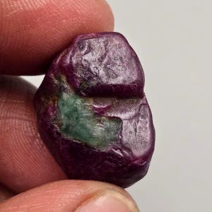 Pode incluir: Uma pedra preciosa bruta e não polida com uma paleta de cores magenta e roxa profunda e saturada, com uma proeminente inclusão verde-azulado opaca incrustada no centro da pedra, exibindo uma superfície natural, irregular e texturizada.