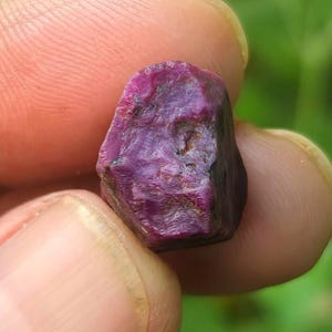 Pode incluir: Uma pedra preciosa bruta e não polida com um tom magenta e roxo profundo. A pedra apresenta uma superfície texturizada e irregular com estruturas cristalinas visíveis e inclusões escuras e opacas em um dos lados.