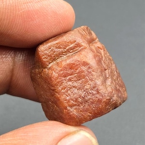Puede incluir: Una piedra preciosa en bruto y sin pulir con una forma irregular y compacta, de color marrón rojizo intenso. La superficie de la piedra muestra una apariencia cristalina y texturizada con facetas visibles y surcos naturales en su estructura.