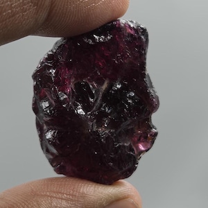 Peut inclure: Une pierre précieuse grenat brute d'un rouge profond avec une texture de surface irrégulière et facettée. La pierre présente des qualités translucides avec des tons cramoisi foncé et des bords tranchants sur toute sa structure.