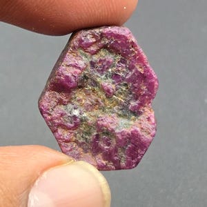 May include: A raw, hexagonal-shaped ruby gemstone featuring a deep magenta and crimson colour palette with visible natural inclusions, textured surface variations, and earthy dark grey mineral deposits embedded within the crystalline structure.