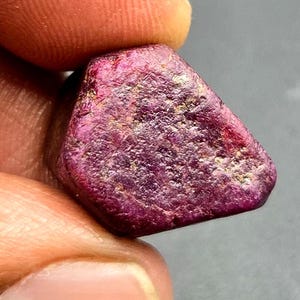 Puede incluir: Una piedra preciosa de rubí en bruto y sin tallar de color magenta intenso y rojo oscuro. La piedra tiene una superficie rugosa y texturizada con facetas cristalinas visibles e inclusiones naturales, con una forma irregular y redondeada.