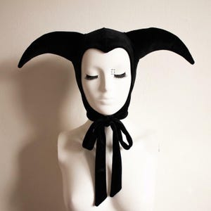 May include: A black velvet hood featuring two long, pointed, horn-like structures extending from the sides. The headpiece has a fitted design that frames the face with a pointed forehead detail and secures under the chin with two long fabric ties.