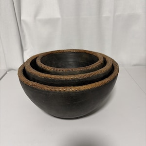 May include: A set of three nested dark gray bowls with smooth, matte surfaces. Each bowl features a distinct, textured rim crafted from woven brown natural fibers, creating a contrasting border around the circular opening of each individual vessel.