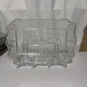 May include: A rectangular clear glass serving tray featuring a molded relief of The Last Supper in the center. The border is decorated with a raised pattern of grape clusters and leaves. The tray has a scalloped edge and a smooth, glossy finish.